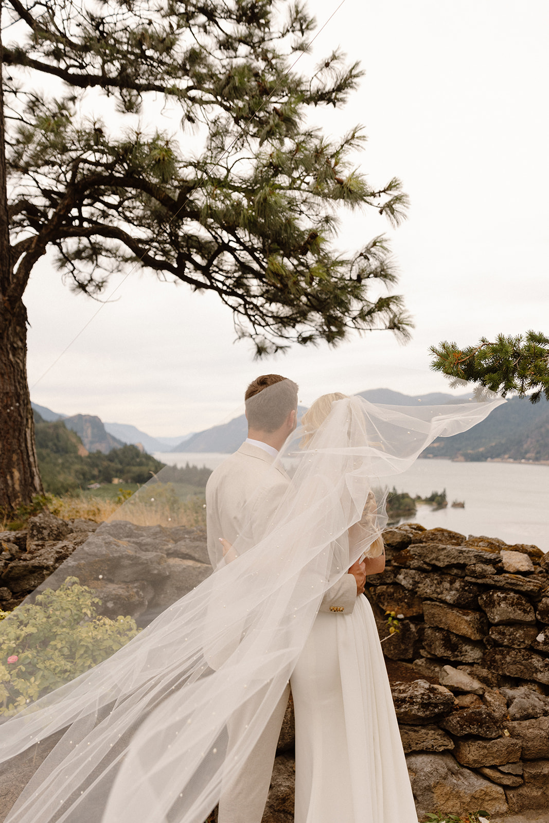 Brides veil blowing in the wind as the groom hold her while they look out at the Columbia river gorge