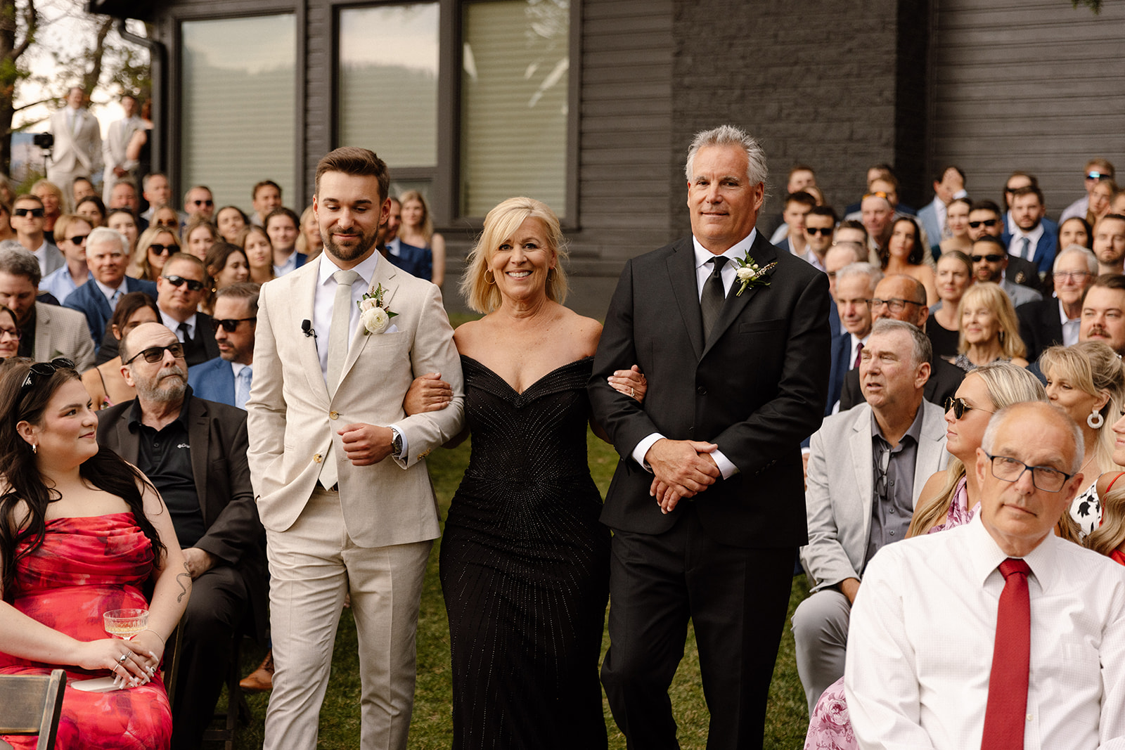 groom walking down the aisle with him mom and dad surrounded by family and friends and lush greenery